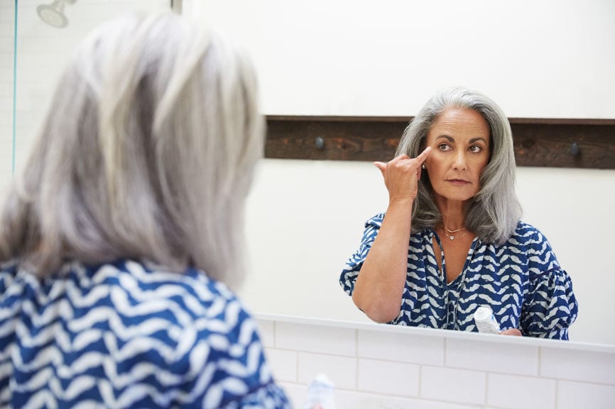 Senior Woman With Grey Hair Putting On Moisturizer In Bathroom Mirror